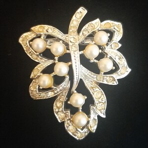 May include: A silver-toned leaf-shaped brooch with faux pearls and clear rhinestones. The brooch features a central vein with pearl accents and rhinestone-studded edges, creating a sparkling, elegant accessory.
