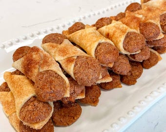 May include: A platter of rolled pastries, each featuring a light-colored, golden-brown dough shell dusted with cinnamon sugar, wrapped around a dense, textured, round brown cookie center. The pastries are arranged in a layered, overlapping stack.
