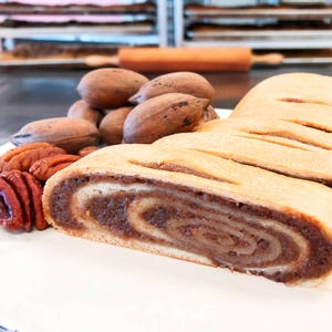 May include: A sliced nut roll pastry with a golden-brown crust and a dense, swirled brown filling, accompanied by several whole and shelled pecans on a white surface. The pastry shows a distinct spiral pattern of dough and nut filling.