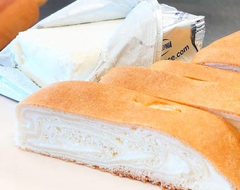 May include: A sliced golden-brown pastry roll with a visible white cream cheese swirl filling, positioned next to an unwrapped block of Philadelphia cream cheese in its silver foil packaging and a wooden rolling pin handle in the background.