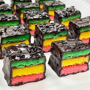 May include: A collection of square-shaped petit fours arranged in rows. Each treat features three distinct horizontal layers of cake in green, yellow, and pink, fully coated in a dark chocolate glaze and topped with small dark chocolate sprinkles.