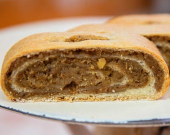 May include: A slice of nut roll pastry featuring a golden-brown baked crust and a dense, spiraled interior filling made of finely ground nuts. The pastry is positioned horizontally, showcasing the layered texture of the dough and the nut mixture.