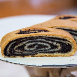 May include: A slice of poppy seed roll featuring a golden-brown baked exterior and a dense, spiraled interior filling of dark, finely ground poppy seeds layered within a light-colored dough, presented on a white surface with a dark rim.