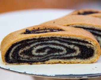 May include: A slice of poppy seed roll featuring a golden-brown baked exterior and a dense, spiraled interior filling of dark, finely ground poppy seeds layered within a light-colored dough, presented on a white surface with a dark rim.