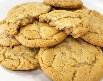 May include: A pile of golden-brown, round cookies with a cracked, textured surface. The cookies are baked to a light tan color, showing soft, chewy centers and slightly crisp edges. They are stacked together on a white plate with a scalloped edge.