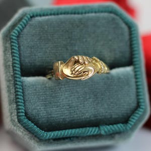 May include: A gold-colored ring featuring a design of two hands clasped together, set in a teal velvet ring box. Other velvet ring boxes in pink and red are visible in the background.