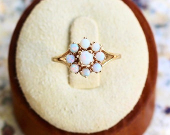 May include: A gold ring featuring a cluster of opals arranged in a floral design. The ring is set on a cream-colored velvet surface inside a brown jewelry box. The opals are white with a subtle play of color.