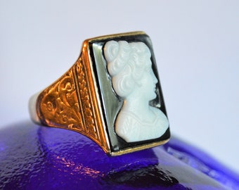 May include: A gold ring with a black and white cameo featuring a woman's profile. The ring has an ornate design on the band.