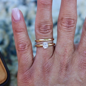 May include: A close-up of a hand wearing a gold wedding band and an oval-shaped diamond engagement ring.