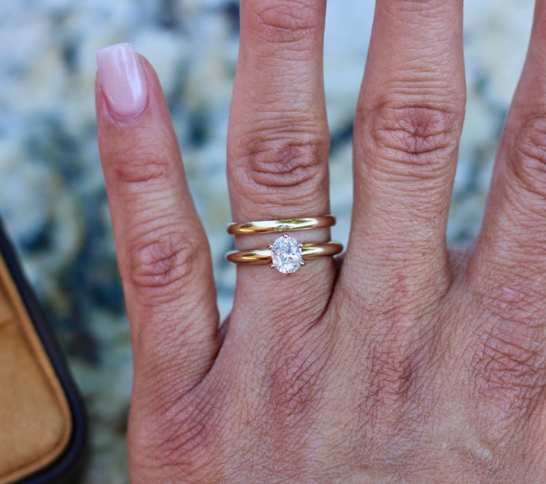 May include: A close-up of a hand wearing a gold wedding band and an oval-shaped diamond engagement ring.