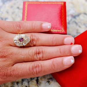 May include: A pear-shaped ruby ring with a halo of baguette-cut diamonds set in yellow gold. The ring is on a hand with white nail polish and small white heart designs.