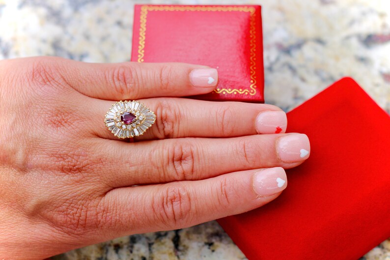 May include: A pear-shaped ruby ring with a halo of baguette-cut diamonds set in yellow gold. The ring is on a hand with white nail polish and small white heart designs.