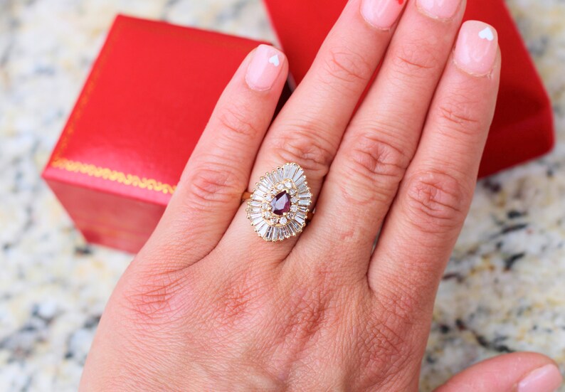 May include: A gold ring with a pear-shaped red gemstone surrounded by smaller white gemstones.