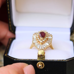 May include: A gold ring with a pear-shaped ruby surrounded by a halo of smaller diamonds. The ring is in a black ring box with a white interior.