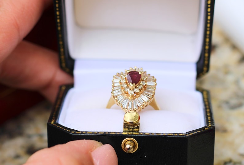May include: A gold ring with a pear-shaped ruby surrounded by a halo of smaller diamonds. The ring is in a black ring box with a white interior.