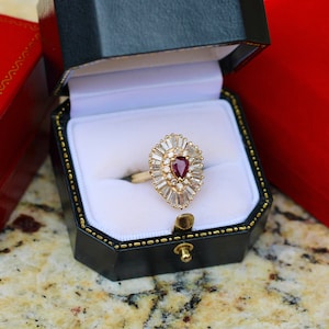 May include: A pear-shaped ruby ring set in a gold band with a halo of baguette-cut diamonds. The ring is in a black and white ring box.