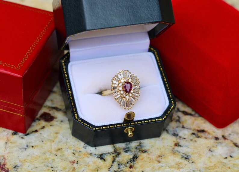 May include: A pear-shaped ruby ring set in a gold band with a halo of baguette-cut diamonds. The ring is in a black and white ring box.