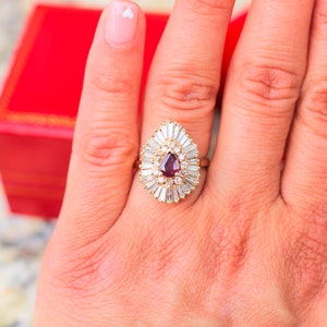 May include: A pear-shaped ruby ring with a halo of baguette-cut diamonds set in yellow gold.