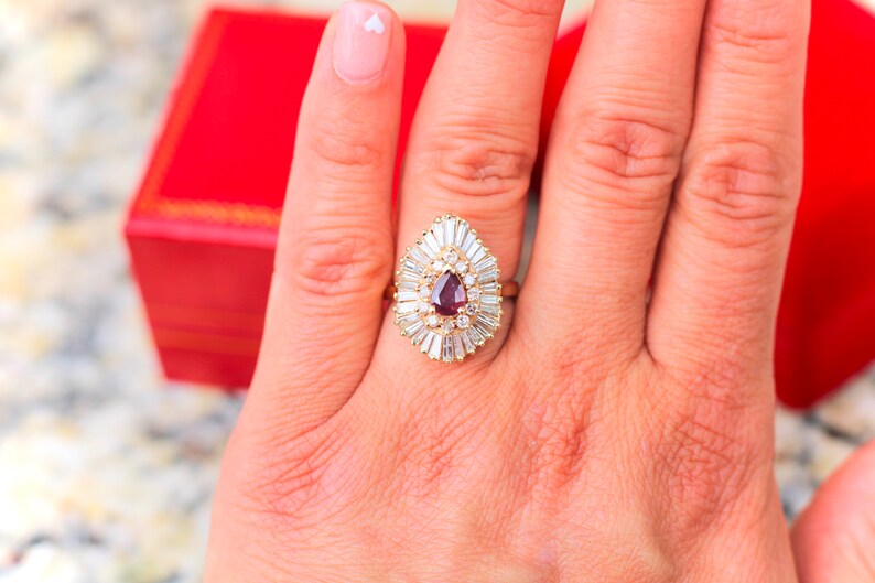 May include: A pear-shaped ruby ring with a halo of baguette-cut diamonds set in yellow gold.