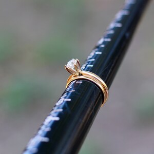 May include: A gold engagement ring with a single, round, clear diamond set in a prong setting. The ring is on a black measuring tool.
