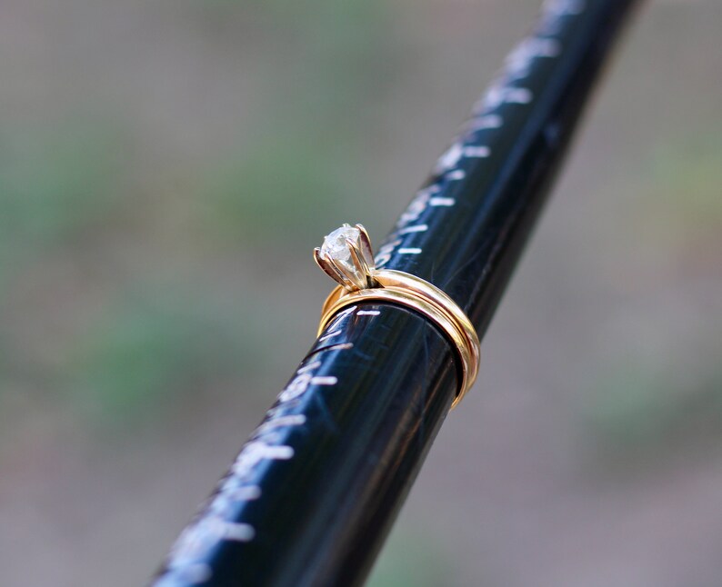 May include: A gold engagement ring with a single, round, clear diamond set in a prong setting. The ring is on a black measuring tool.