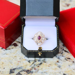 May include: A red ruby ring with a pear-shaped stone surrounded by smaller diamonds, set in a gold band. The ring is displayed in a black ring box with a white interior.