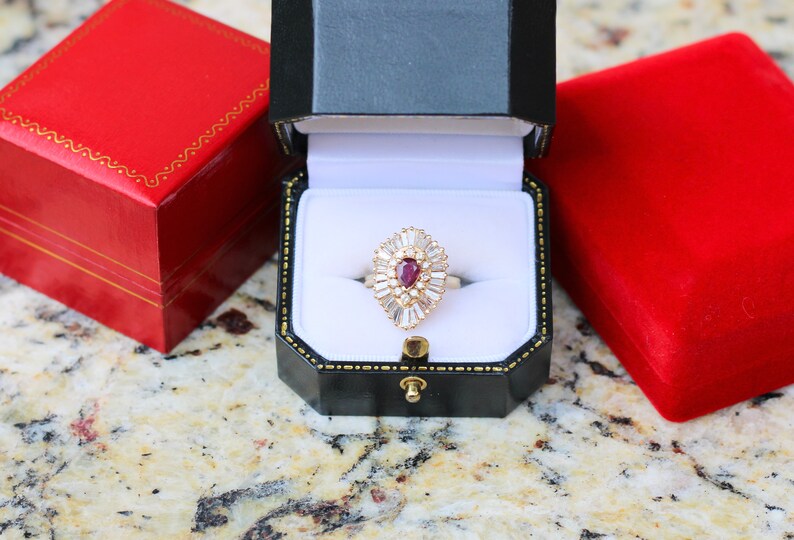 May include: A red ruby ring with a pear-shaped stone surrounded by smaller diamonds, set in a gold band. The ring is displayed in a black ring box with a white interior.