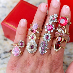 May include: A hand wearing a variety of rings with pink and red gemstones. The rings are set in gold and silver and feature different styles, including pear-shaped, round, and cluster designs.