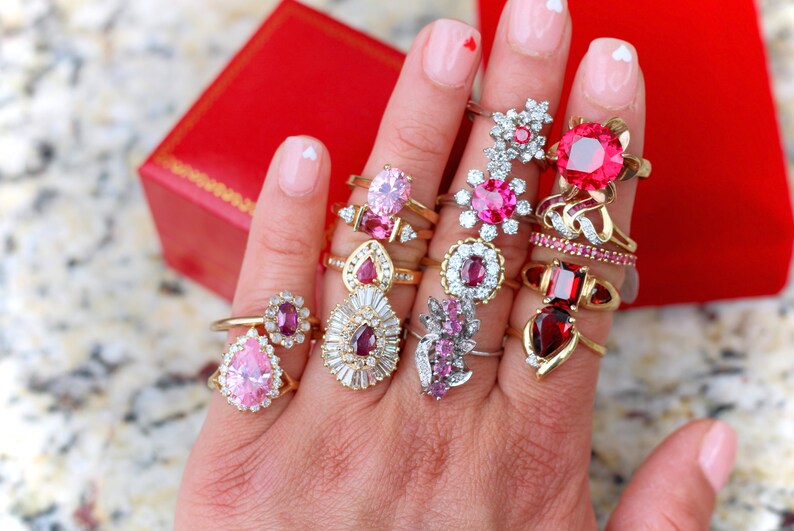 May include: A hand wearing a variety of rings with pink and red gemstones. The rings are set in gold and silver and feature different styles, including pear-shaped, round, and cluster designs.
