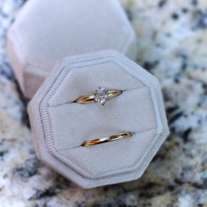 May include: Two gold rings in a white velvet ring box. The top ring has a large, clear, round diamond. The bottom ring is a plain band.