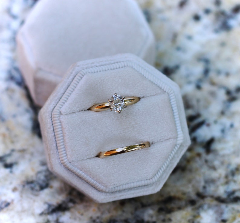 May include: Two gold rings in a white velvet ring box. The top ring has a large, clear, round diamond. The bottom ring is a plain band.