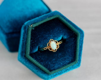 May include: A gold ring with an oval opal stone set in a delicate filigree design, displayed in a teal blue velvet ring box.