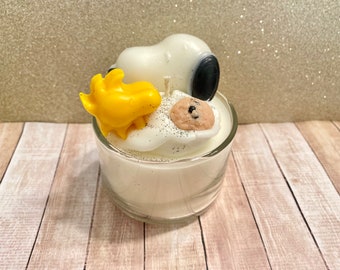 May include: A white candle in a glass jar with a yellow Snoopy figurine, a white Snoopy figurine with a black nose, and a chocolate chip cookie figurine on top.
