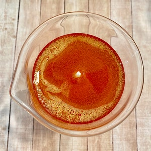 May include: A clear glass beaker filled with a bright orange liquid. The liquid has a slight swirl pattern and small bubbles throughout.