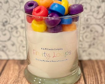 May include: A glass jar candle with a white label that reads "A & M Candle Company Fruity Loops 12 oz. Soy Mix Wax & Made with Love in N.C.". The candle is topped with colorful, fruity-shaped wax melts.
