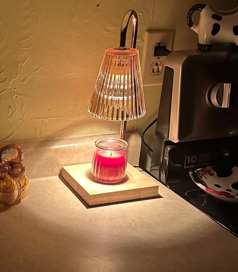 May include: A glass candle warmer with a clear glass shade and a wooden base. A red candle is placed on the base. The warmer is plugged into an electrical outlet.