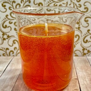 May include: A clear glass beaker filled with orange gel candle with a white wick.