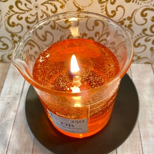 May include: A clear glass beaker candle with a flickering flame. The candle is filled with a shimmering orange wax and has a label that reads "250" and "Orange Blossoms".