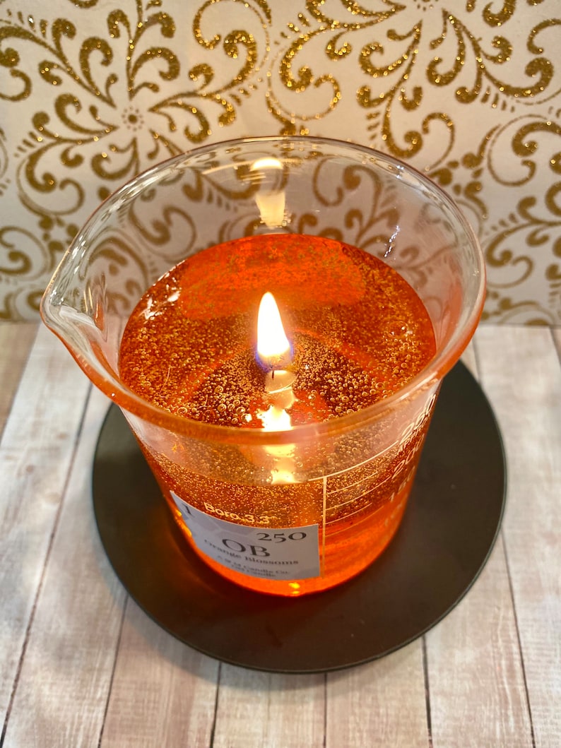 May include: A clear glass beaker candle with a flickering flame. The candle is filled with a shimmering orange wax and has a label that reads "250" and "Orange Blossoms".