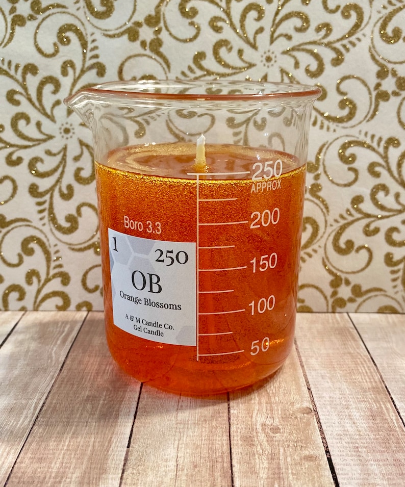 May include: A clear glass beaker filled with orange gel candle wax. The beaker has markings for measuring volume in milliliters. The label on the beaker reads "1, Boro 3.3, 250, OB, Orange Blossoms, A & M Candle Co., Gel Candle".