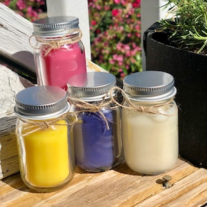 Pode incluir: Quatro pequenos frascos de vidro com tampas prateadas e detalhes de corda cheios de velas coloridas. As velas são amarelas, roxas, brancas e rosas.