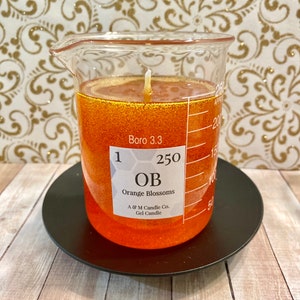 May include: A clear glass beaker filled with orange gel candle with a white wick. The beaker has a label that reads "1 250 OB Orange Blossoms A & M Candle Co. Gel Candle" and "Boro 3.3". The beaker is sitting on a black circular base.