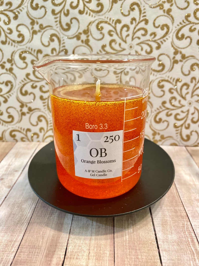 May include: A clear glass beaker filled with orange gel candle with a white wick. The beaker has a label that reads "1 250 OB Orange Blossoms A & M Candle Co. Gel Candle" and "Boro 3.3". The beaker is sitting on a black circular base.