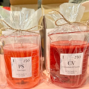 May include: Two clear glass beakers filled with red gel candles. The beaker on the left is labeled "PS Pink Sugar" and the beaker on the right is labeled "CV Cinnamon & Vanilla". Both beakers are labeled with "1 250" and "A & M Candle Co. Gel Candle".