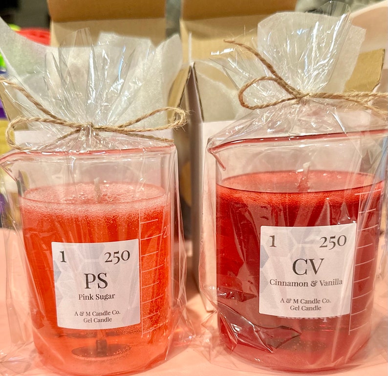May include: Two clear glass beakers filled with red gel candles. The beaker on the left is labeled "PS Pink Sugar" and the beaker on the right is labeled "CV Cinnamon & Vanilla". Both beakers are labeled with "1 250" and "A & M Candle Co. Gel Candle".