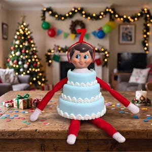 May include: A doll with a brown-haired head and red limbs wearing a light blue, three-tiered cake costume with white scalloped trim. A red headband with the number 7 sits on the doll's head, and the doll is positioned on a wooden table.