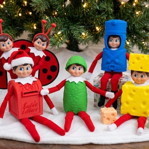May include: Five elf dolls in various costumes: two as ladybugs, one in a red mailbox suit labeled 'Santa's Letters', one in a green pickle suit, one in a blue toy soldier outfit, and one in a yellow Swiss cheese block suit.