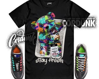 Peut inclure: T-shirt noir avec un motif d'ours en peluche coloré dans un emballage plastique et le texte "stay fresh". L'image montre également une paire de baskets noires et grises avec des détails colorés.