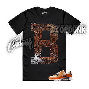 May include: Black t-shirt with a large letter "B" filled with the word "Blessed" in various sizes and shades of brown. The text "This T Shirt Was Made and Designed by Cordunk" is printed on the shirt. A pair of orange and tan sneakers are at the bottom.