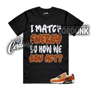 May include: Black t-shirt with the text "I MATCH ENERGY SO HOW WE GON ACT?" in white and orange. The shirt is paired with orange and tan sneakers. The brand name "Cordunk" is visible.